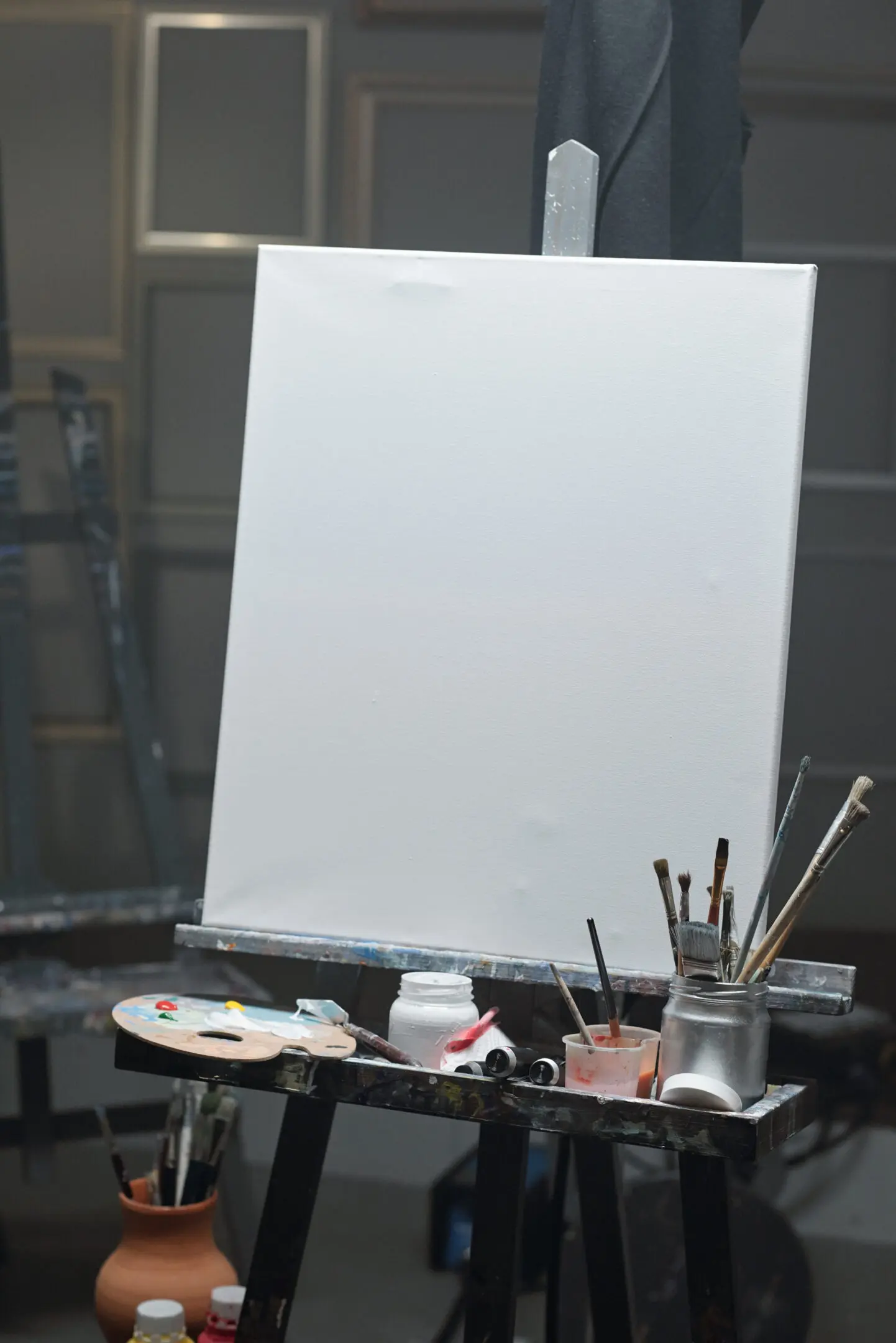 Blank sheet of paper on easel with color palette, jars and jug with paintbrushes and other painting stuff near by