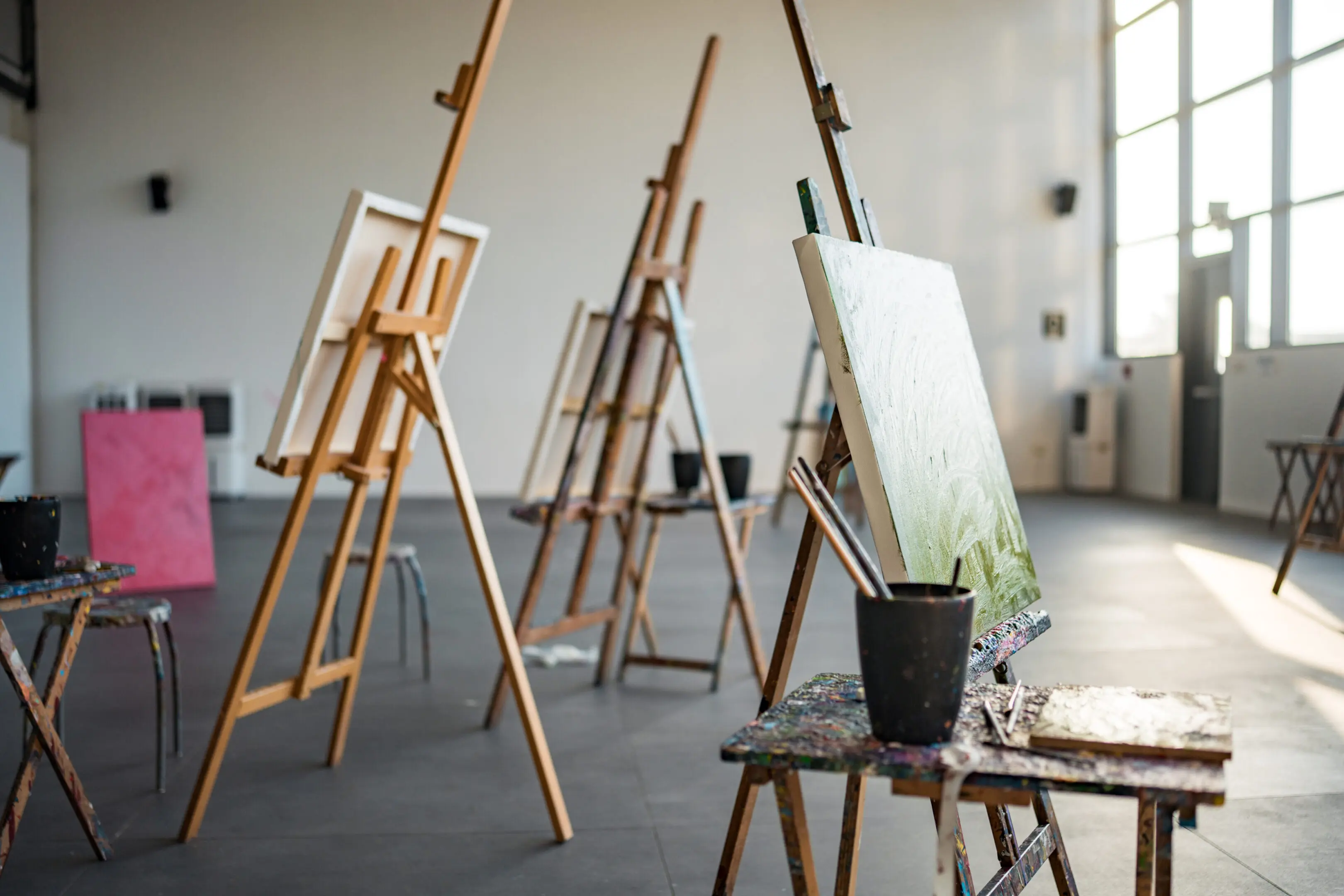 A spacious art studio with vibrant easels and canvas stands, bathed in natural light from large windows. Paint supplies are neatly arranged, creating an inspiring atmosphere for creativity and artistic expression.